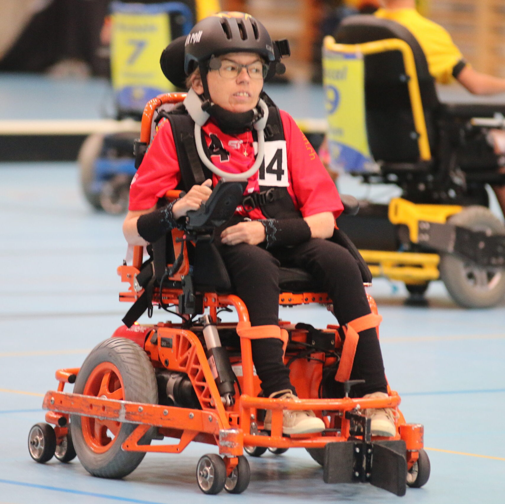 Matchsituation: Simone Leuenberger mit schwarzem Helm, Halskrause, rotem Matchshirt mit Nummer 14 im orangen Sportrollstuhl,, im Hintergrund sind andere Spieler und die Bande verschwommen sichtbar.