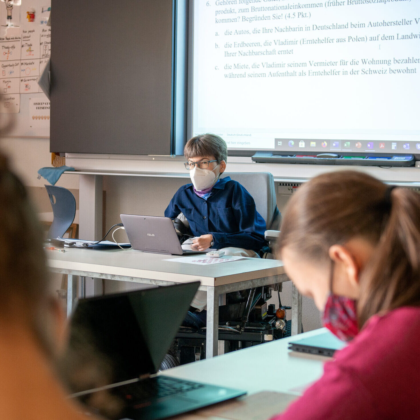 Im Vordergrund drei Schülerinnen von hinten an Pulten, im Hintergrund Simone Leuenberger am Lehrerpult, dahinter Wandtafel und Leinwand mit Text