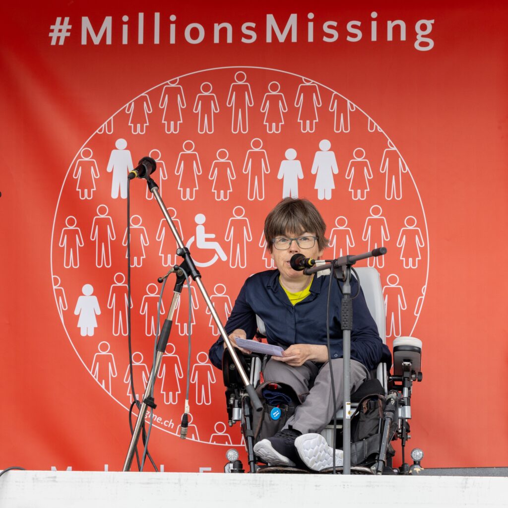 Simone Leuenberger hinter einem Mikrofon, dahinter das Logo von MillionsMissing: Weisse, schematisch dargestellte Menschen mit und ohne Rollstuhl auf rotem Grund.