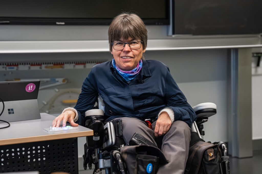 Simone Leuenberger im Elektrorollstuhl neben dem auf Sitzhöhe heruntergestellten Lehrpersonenpult. Sie hat die rechte Hand auf Knöpfen auf dem Pult. Auf dem Pult steht ein Laptop. Im Hintergrund sieht man eine Wandtafel.